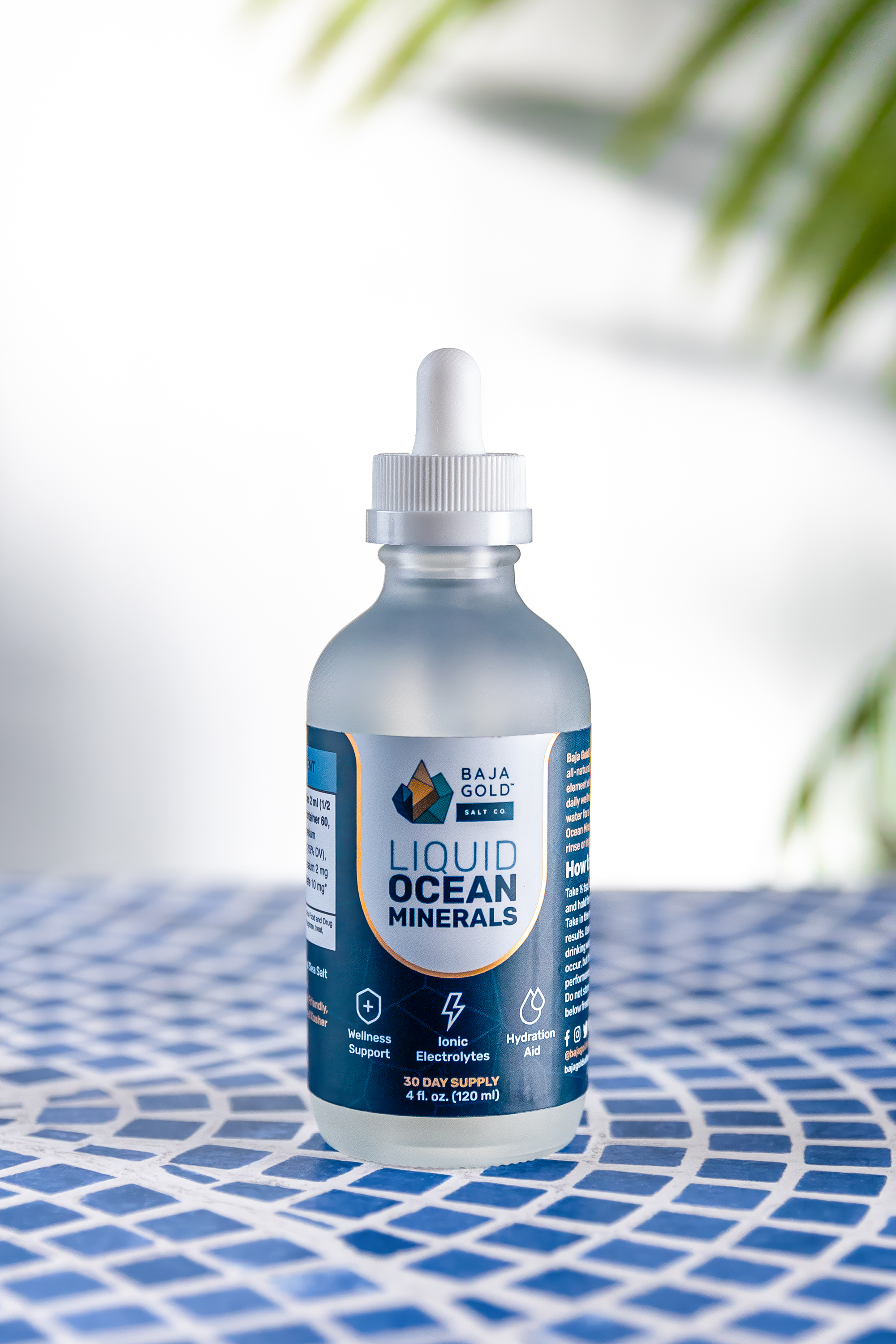 Bottle of Baja Gold liquid ocean minerals on a blue and white patterned surface with blurred greenery in the background.