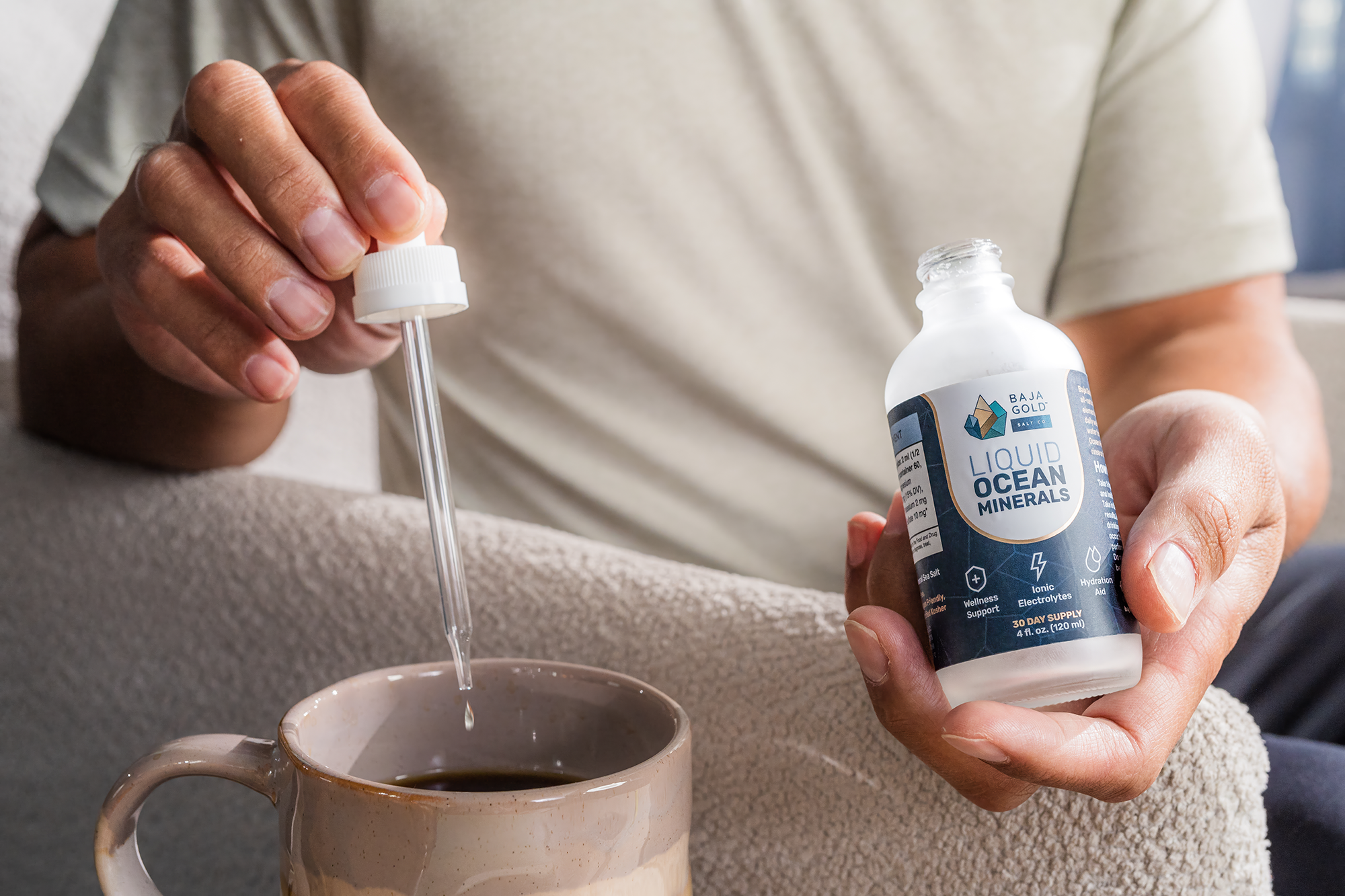 Person adding drops from a bottle labeled 'Baja Gold liquid ocean minerals' into a cup of coffee.
