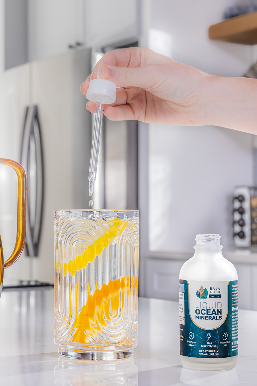 Person adding drops of Baja Gold liquid ocean minerals to a glass of water in a kitchen setting.