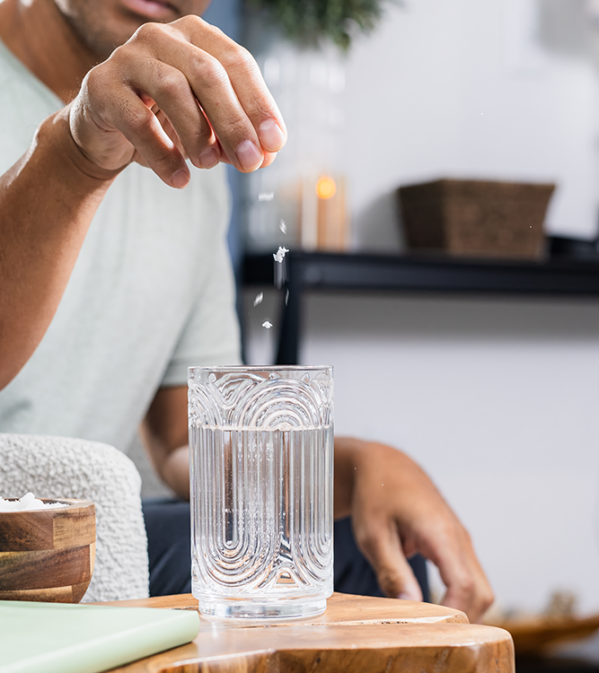 Person sprinkling natural grain Baja Gold mineral sea salt into a glass of water on a wooden table.